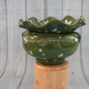 May include: A glossy olive green ceramic planter with a ruffled, wavy rim and a speckled finish featuring white and dark blue spots. The bowl-shaped vessel sits atop a cylindrical light-colored wooden base, showcasing a smooth, glazed texture.