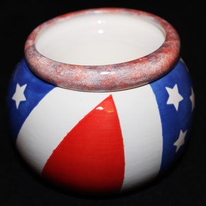 May include: A white ceramic planter with a red, white, and blue American flag design. The planter has a wide rim with a speckled red and blue glaze.
