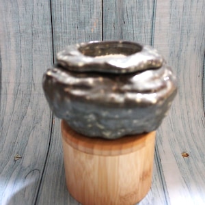 May include: A metallic, dark gray, unevenly shaped ceramic bowl with a glossy finish, resting on a cylindrical light-colored wooden base. The bowl features a thick, textured rim and a deep, rounded interior cavity for holding small items.