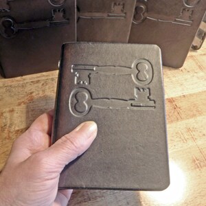 May include: A dark brown rectangular notebook with a smooth, matte finish. The front cover features an embossed design of two vintage-style skeleton keys positioned horizontally, one above the other, facing in opposite directions.