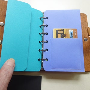 May include: A brown leather bound notebook with a turquoise blue and a purple pocket on the inside. The notebook has a metal ring binder and a brown leather strap.