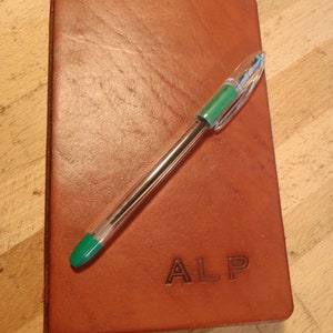 May include: A brown leather journal with the initials "ALP" embossed on the front cover. A green and clear pen is lying on top of the journal.