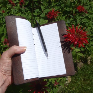 May include: A brown leather bound journal with a black pen lying on the open pages. The journal is being held by a hand in front of a background of red flowers.