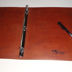 May include: A brown leather binder with a silver three-ring mechanism. The binder has a black logo that says "Sherwood Leather". A black pen is lying on the binder.