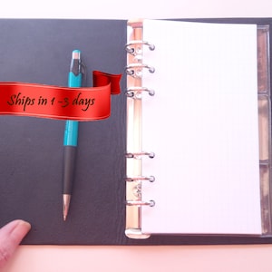 May include: A black leather bound planner with a silver ring binder and a blue pen. The planner has a clear plastic pocket on the right side and a red ribbon banner that says "Ships in 1-3 days".