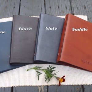 May include: Four leather-bound binders in different colors: blue, black, slate, and saddle. The binders are arranged in a row from left to right. The saddle binder has the word "RECIPES" embossed on the front.
