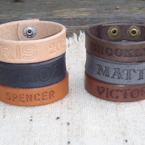 May include: Four leather bracelets stacked on top of each other. The top bracelet is light brown and says "PARIS 2021". The next bracelet is black and says "ANTHONY". The third bracelet is brown and says "SPENCER". The bottom bracelet is brown and says "VICTOR".