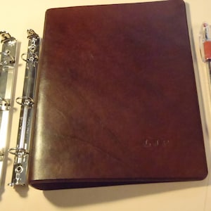 May include: A brown leather binder with silver metal rings and a silver metal spine. The binder is closed and has a subtle embossed design.