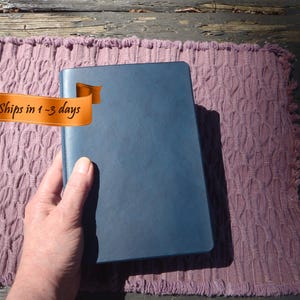 May include: A blue notebook with a smooth cover is held over a textured, mauve-colored mat. The notebook is rectangular and appears to be closed. An orange banner with the text "Ships in 1-3 days" is in the image.