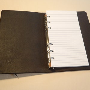 May include: A black leather bound notebook with a silver ring binder and lined paper. The notebook is open to a blank page.