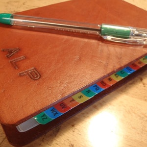 May include: A brown leather bound journal with the word "ALP" embossed on the cover. The journal has colorful tabs along the edge, labeled with letters of the alphabet.