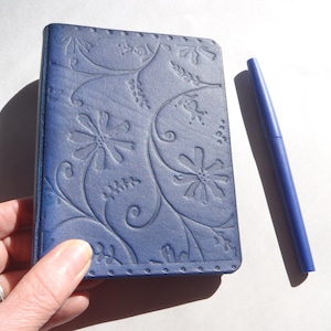 May include: A rectangular navy blue notebook with a debossed floral pattern on the cover, featuring stylized flowers and swirling vines. The notebook has a stitched border detail and is accompanied by a matching blue ballpoint pen.