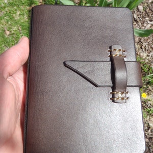 May include: A dark brown leather journal featuring a smooth, rectangular cover with rounded corners. A horizontal leather strap closure is secured to the front cover with two sets of light-colored cross-stitching, creating a simple, functional design.