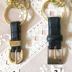 May include: Two gold keychains with black leather straps and gold buckles. The keychains have multiple gold rings attached to them. The keychains are on a white lace background.