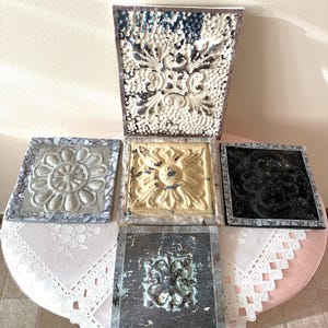 May include: An assortment of decorative metal tiles with embossed floral and geometric designs. The tiles feature various colors, including white, black, silver, and yellow, with distressed finishes. The tiles are arranged on a pink table with a lace tablecloth.