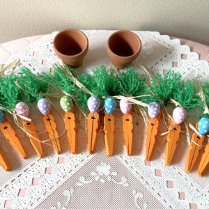 May include: A set of 12 carrot-shaped clothespins decorated with green and blue Easter egg designs. The clothespins are arranged on a white lace tablecloth with two small terracotta pots in the background.