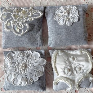 May include: Four gray fabric sachets with white lace and pearl embellishments. The sachets are square and have a soft, textured surface.