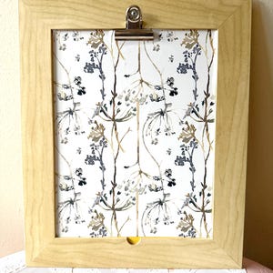 May include: A light wood picture frame with a clip at the top. The frame holds a print with a white background and a pattern of brown and gray stylized flowers.