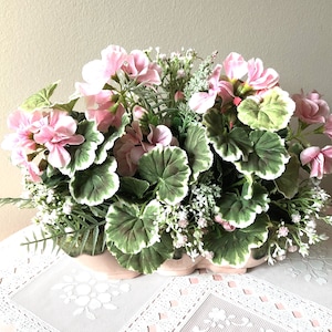 May include: A pink and green artificial flower arrangement in a pink ceramic planter. The arrangement features pink geraniums and green leaves with white edges.