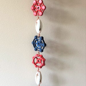 May include: A red, white, and blue hanging mobile made from repurposed metal water valve handles. The mobile features a variety of shapes and sizes of the handles, with some painted red, some painted blue, and some left in their natural metal finish. The mobile is suspended from a copper wire loop.