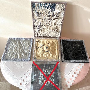 May include: A collection of four square metal ceiling tiles with embossed patterns. One tile is large and rectangular with white and dark distressed paint, while the others are square with silver, gold, and black finishes. One tile is marked with a red X.
