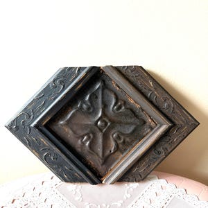 May include: A diamond-shaped, decorative wall panel with a dark, distressed finish. The panel features a central embossed design and a detailed, carved border. The color palette includes shades of black, brown, and gray, creating a rustic aesthetic.