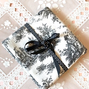 May include: A white fabric gift bag with a black and white toile pattern. The bag is tied with a black ribbon with white polka dots.