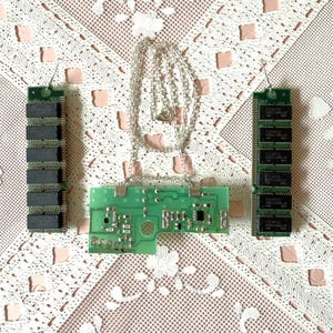 May include: A green circuit board with a silver chain necklace, two green and black computer memory chips, and a white lace tablecloth.