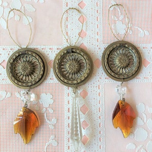 May include: Three gold-toned metal ornaments with a floral design and a crystal drop. The ornaments are hanging from a wire loop and are on a white lace background.