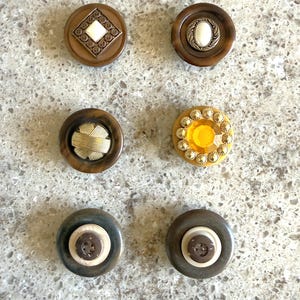 May include: A collection of six vintage buttons in various designs and colors. The buttons include brown, gold, and yellow tones, with decorative elements such as geometric patterns, faux gems, and textured surfaces. These buttons are ideal for sewing and craft projects.