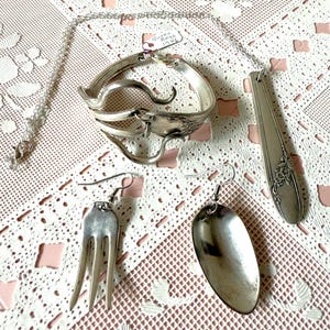 May include: A set of silverware jewelry, including a necklace, bracelet, and earrings. The necklace is a silver chain with a spoon pendant. The bracelet is made from a bent fork. The earrings are a spoon and a fork.
