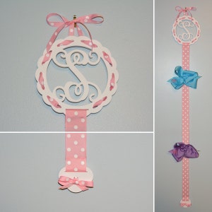May include: White wooden monogram with a pink and white polka dot ribbon hanging from it. The monogram is in the shape of a circle with a scalloped edge. The ribbon is tied in a bow at the bottom.