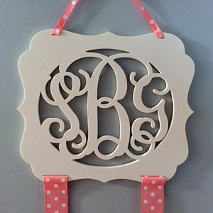 May include: White wooden sign with a cutout monogram of the letters "S B G" in a decorative script font. The sign is hanging from two pink ribbons with white polka dots.