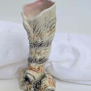 May include: A distinctive ceramic vase with a textured, organic design. The vase has a light beige exterior with brown, black, and green accents. The interior is a soft pink. The base is wide, narrowing to a neck and flared opening.