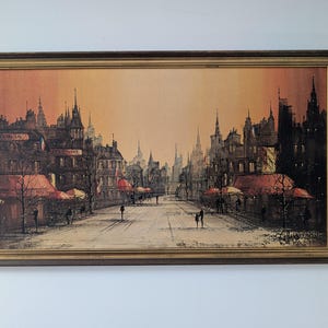 May include: An oil painting depicting a European city street scene, framed in a dark brown wood frame with a gold inner border. The artwork shows buildings, streetlights, and people walking. The sky is a gradient of orange and yellow.