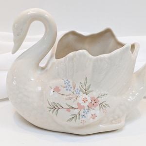 May include: A white ceramic swan planter with a floral design. The planter has a wide opening for planting and a long neck.