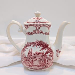 以下が含まれることがあります： 赤い花柄と庭にある家のシーンが描かれた白い陶器製のティーポット。ティーポットには蓋と注ぎ口があります。