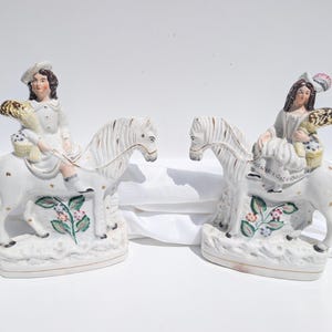 Pode incluir: Duas figuras antigas de porcelana retratando figuras montadas em cavalos brancos. As figuras usam roupas de época e seguram cestas. Os cavalos têm detalhes dourados e florais. As figuras estão em um fundo branco.