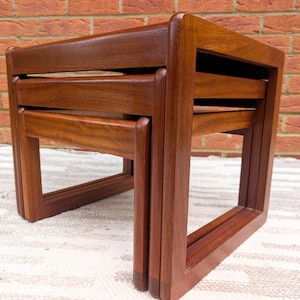May include: A set of three nesting tables in dark brown wood. The tables have a square shape and a simple, geometric design. The wood grain is visible. The tables are stacked together. Each table is approximately 46 cm wide, 46 cm deep, and 41 cm tall.