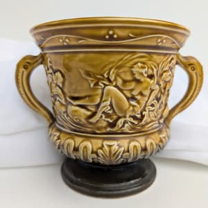 Peut inclure: Un pot en céramique brun avec deux poignées et un motif en relief d'une femme et de feuillage. Le pot a une base brun foncé.