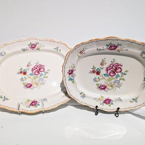 May include: Two oval serving platters with a cream-colored background. Each platter features a floral design with pink, purple, and blue flowers. The edges are scalloped with a gold trim. The platters are displayed on black metal stands.