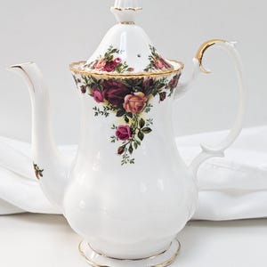 Puede incluir: Tetera de cerámica blanca con un diseño floral de rosas rojas, rosas y moradas. La tetera tiene un ribete dorado en la tapa, el asa y la base. El pico es curvo y la tapa tiene un pequeño adorno.