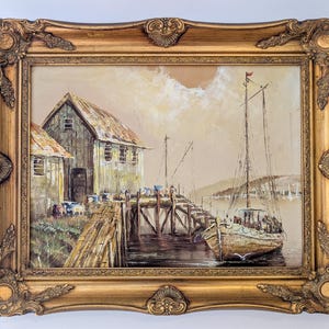 May include: An oil painting in a gold ornate frame depicts a coastal scene. A weathered wooden pier leads to a small boat, with a building on the left. The painting uses muted tones of brown, beige, and gray.
