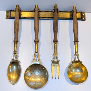 Op de afbeelding: Set van vier gouden keukengerei met houten handvatten die aan een messing rek hangen. De keukengerei omvatten een schuimspaan, een soeplepel, een vork en een lepel.