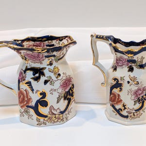 Può includere: Due brocche in ceramica bianca con motivi floreali blu e dorati. La brocca più grande ha un bordo smerlato e un manico. La brocca più piccola ha un bordo dritto e un manico.