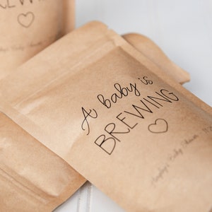 Pu&ograve; includere: Un sacchetto di carta marrone con un cuore bianco e il testo "A baby is BREWING" in corsivo nero.
