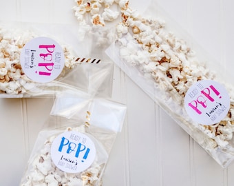 May include: Three clear plastic bags filled with popcorn and tied with gold ribbon. Each bag has a round white sticker with the text "Ready to Pop!" and the name "Lauren's Baby Shower" in black text.