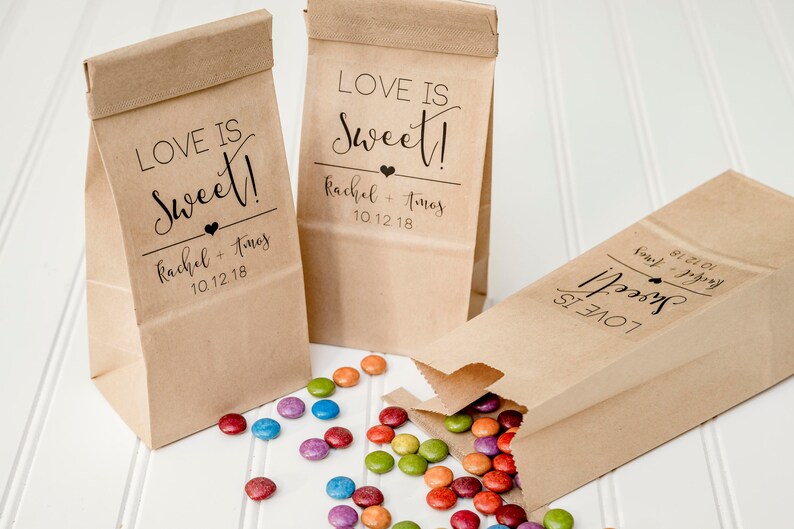 May include: Three brown paper bags with the text "Love is Sweet!" and the names "Rachel + Tomas" and the date "10.12.18". The bags are filled with colorful candy.