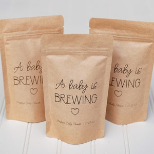 Pu&ograve; includere: Tre sacchetti di carta marrone con un'etichetta bianca su cui &egrave; scritto "A baby is brewing" e un piccolo cuore. I sacchetti sono per una festa di nascita.