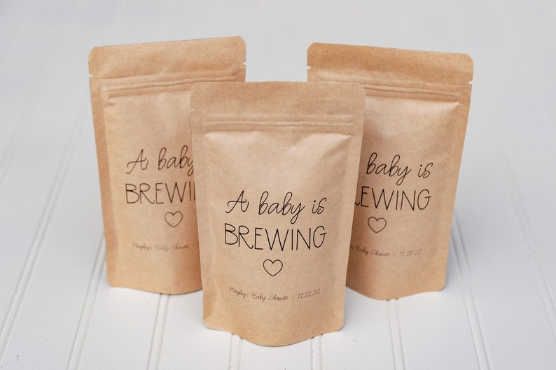 Pu&ograve; includere: Tre sacchetti di carta marrone con un'etichetta bianca su cui &egrave; scritto "A baby is brewing" e un piccolo cuore. I sacchetti sono per una festa di nascita.